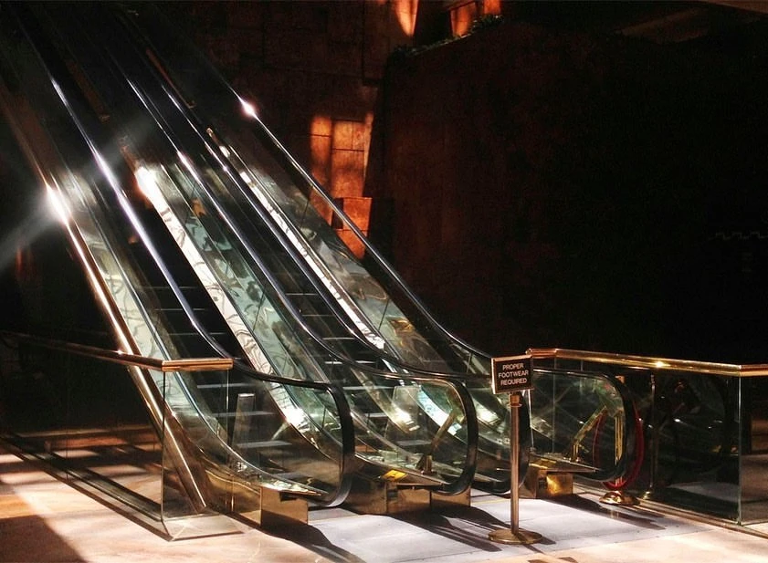 Illuminated escalator with glass railings and metallic steps in an indoor setting, with a sign indicating footwear requirements.