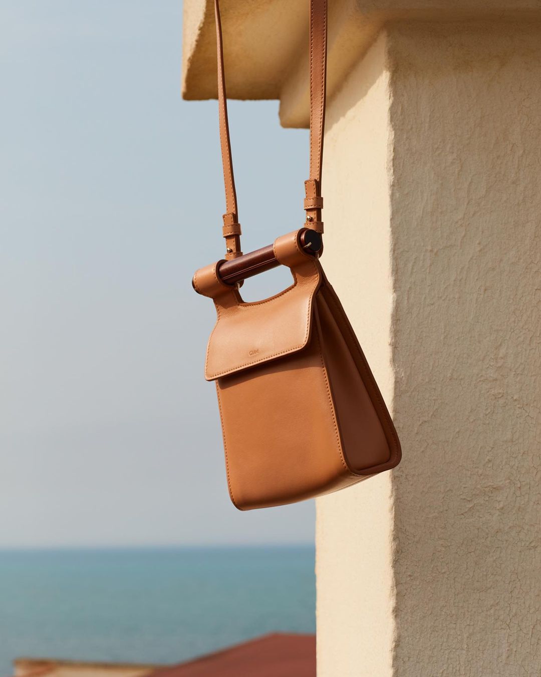 Tan leather purse with wooden handle and shoulder strap photographed outdoors against a blue sea and sky.