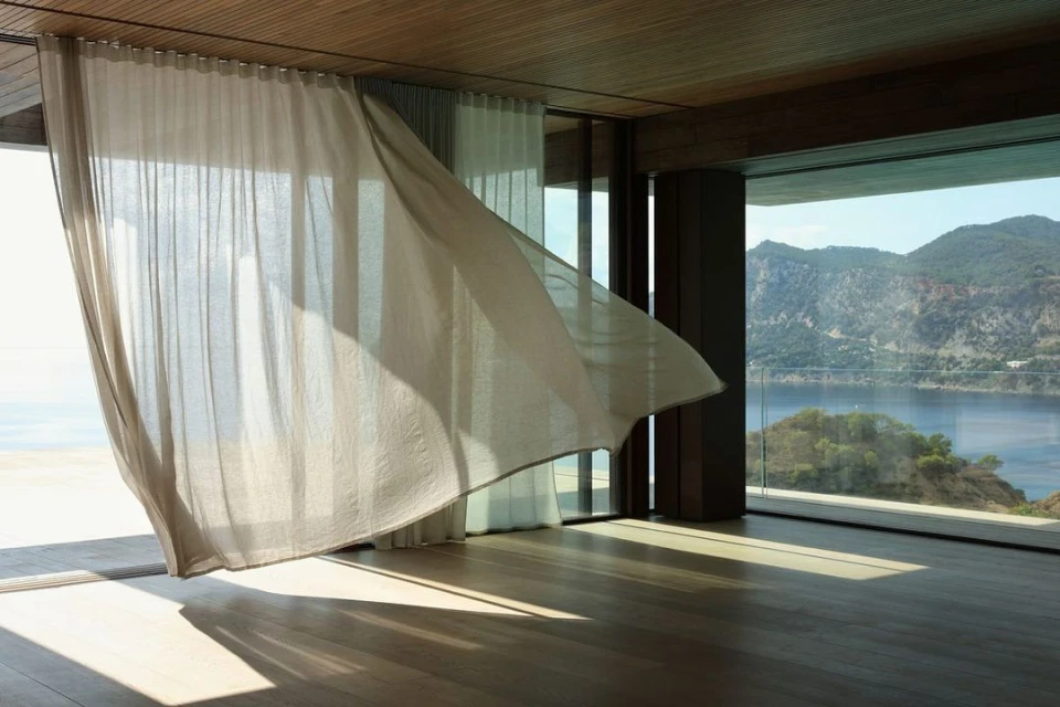 Interior view with a sheer curtain blowing in the breeze and a scenic view of a mountain and ocean through a large window.