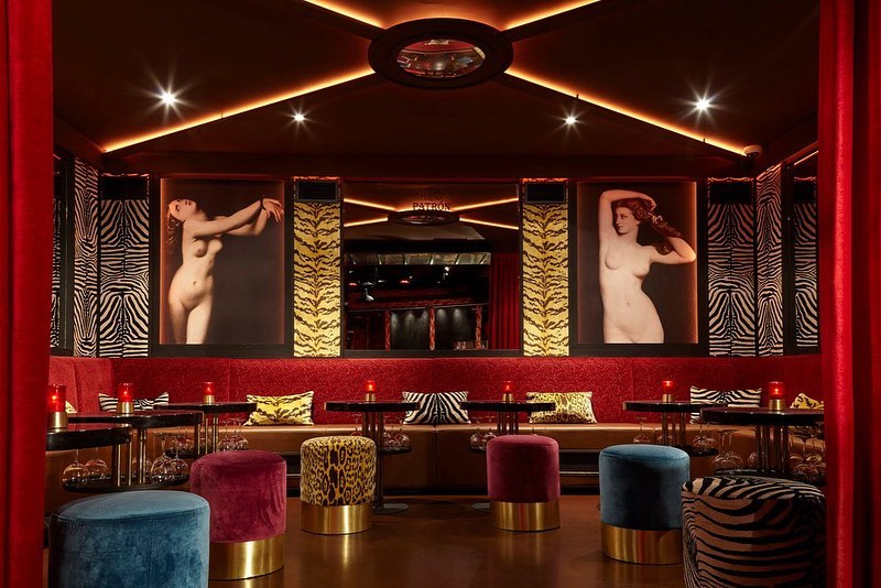 Interior view of a bar with lounge seating and artwork featuring female figures, dimly lit with red and gold accents, creating a warm ambiance.