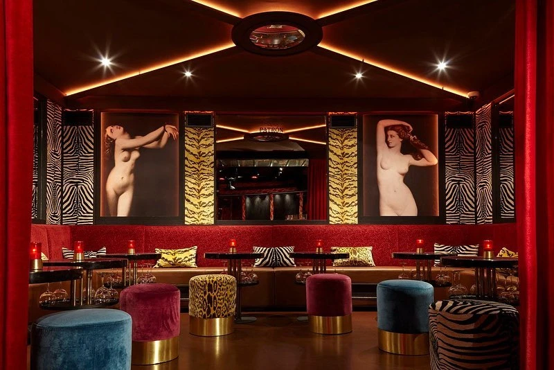 Interior view of a bar with lounge seating and artwork featuring female figures, dimly lit with red and gold accents, creating a warm ambiance.