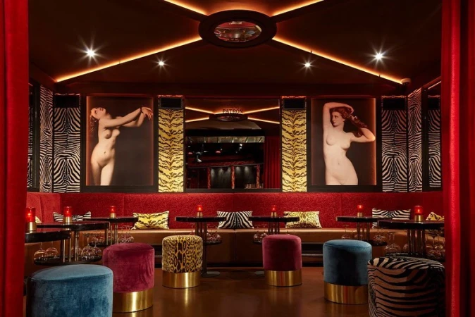 Interior view of a bar with lounge seating and artwork featuring female figures, dimly lit with red and gold accents, creating a warm ambiance.