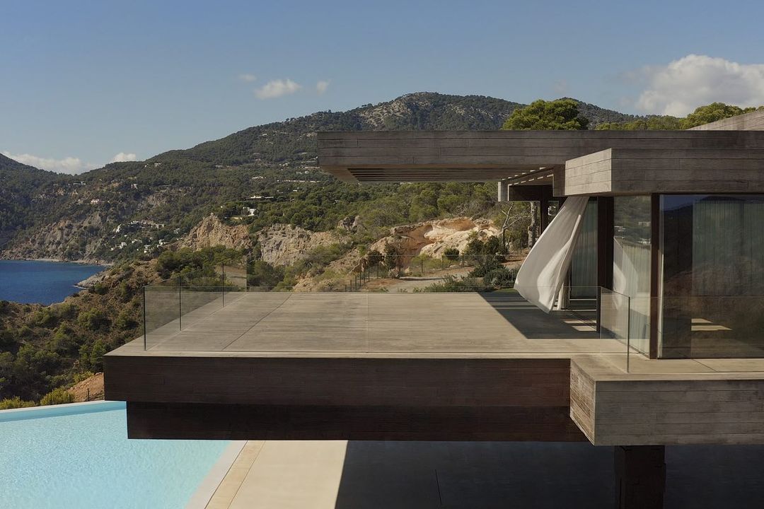 Exterior architecture featuring a modern villa with a deck overlooking an infinity pool, ocean, and mountain landscape under a clear blue sky.