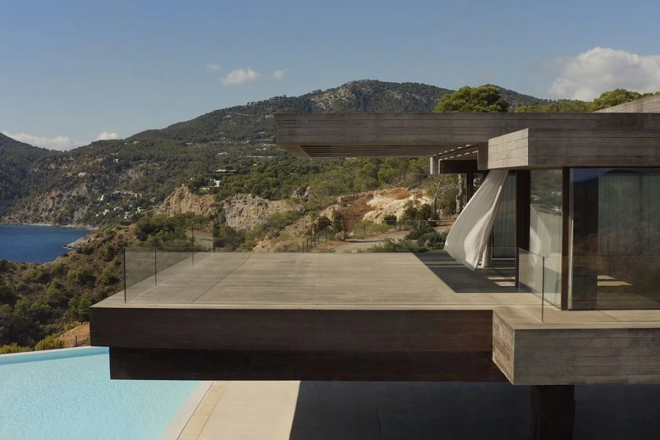 Exterior architecture featuring a modern villa with a deck overlooking an infinity pool, ocean, and mountain landscape under a clear blue sky.
