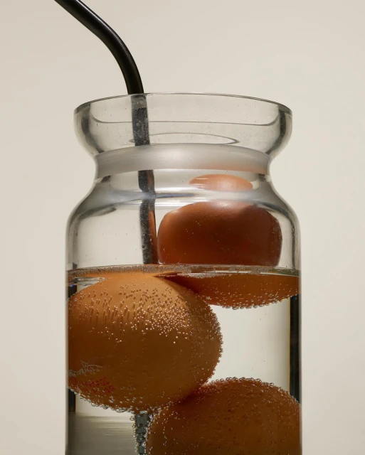 A clear glass jar with three brown eggs suspended in water by a black wire, with bubbles clinging to the eggs.