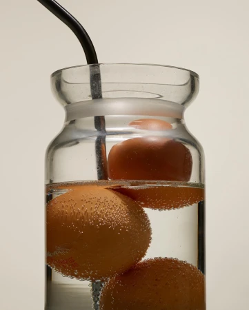A clear glass jar with three brown eggs suspended in water by a black wire, with bubbles clinging to the eggs.
