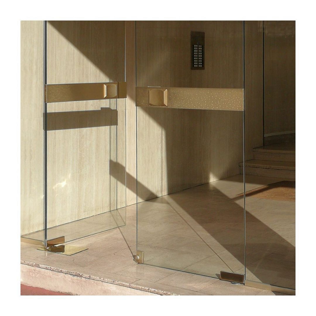 Indoor shot of a glass door with gold hardware details, featuring a push bar, hinges, and frame, next to a light beige stone wall.