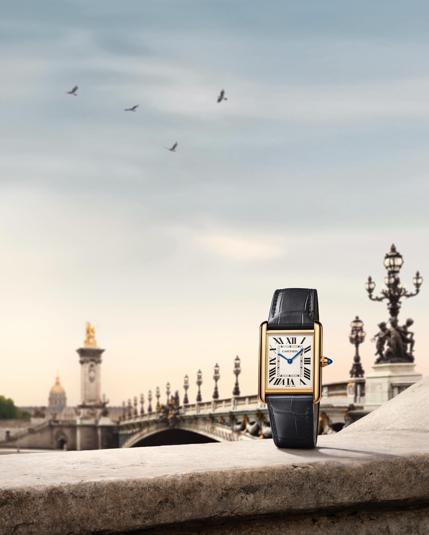 Luxury wristwatch with a leather band is displayed on a stone ledge with a Parisian bridge and the sky in the background.