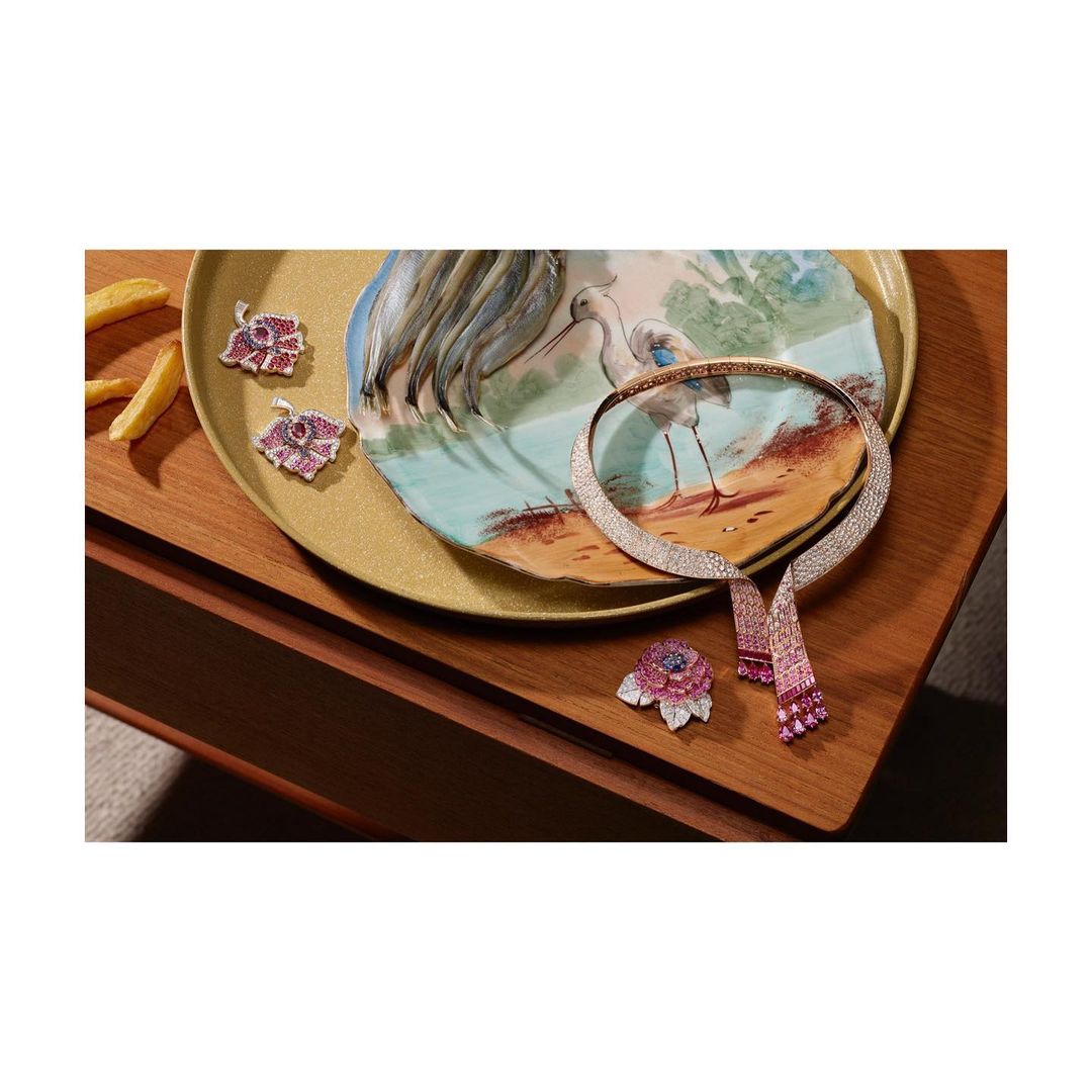 Jewelry still life features a flower brooch, two flower pendants, and a gem-studded choker on a table with a decorative heron plate.