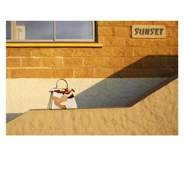 Fashion handbag on a step against a textured tan wall with a sign that reads 'Sunset' in muted, warm lighting creating hard shadows.