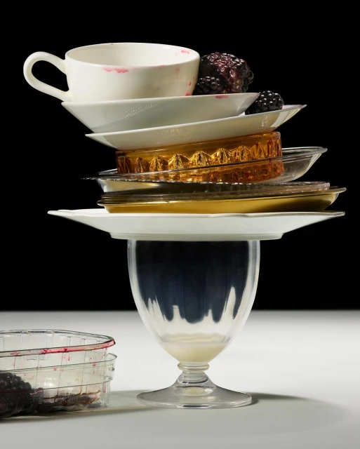 Still life of stacked white and gold plates topped with a teacup with lipstick stain, next to blackberries against black backdrop.