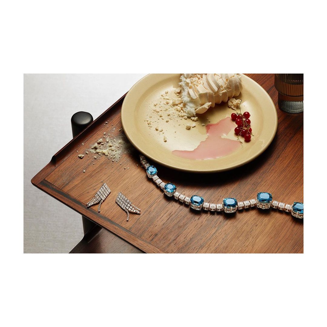 Elevated view showcases diamond earrings, a blue gem necklace, berries, and a dessert on a wooden tray against a light backdrop.