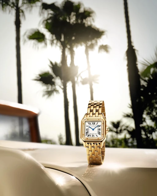 Gold wristwatch with a white face and diamond accents displayed on the hood of a classic car in a sun-drenched tropical setting.