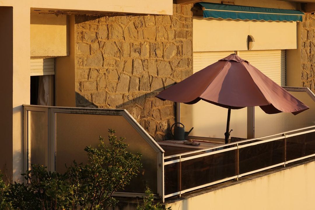 Outdoor patio with a mauve umbrella shading a table and chair on a balcony, greenery visible along the edge of the patio.