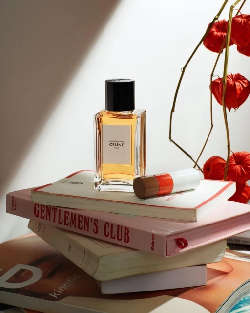Celine perfume bottle stands atop stacked books alongside a brown and white tube. Red seed pods create soft background.