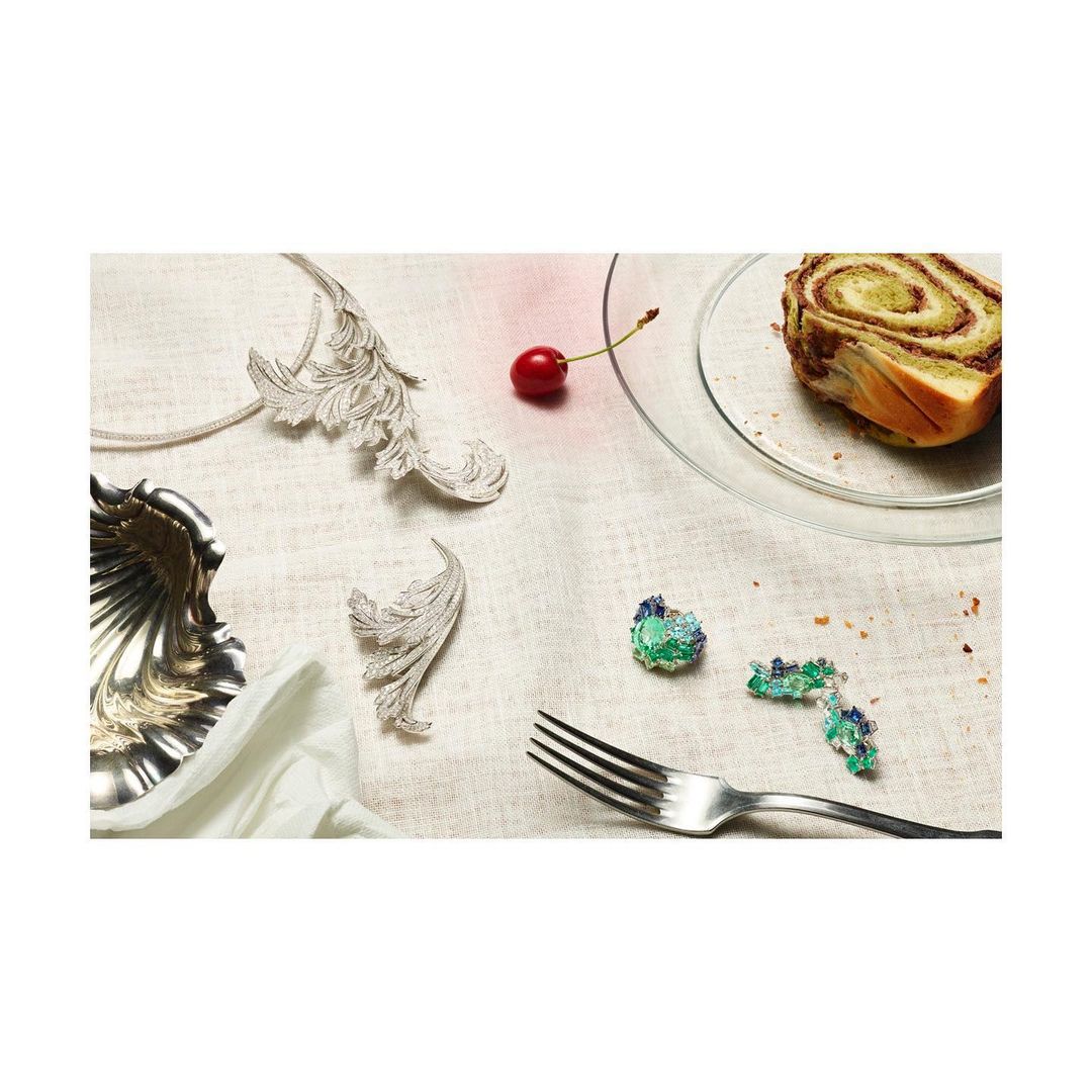 Overhead shot of silver tableware, jewelry, cherry, and pastry on a linen tablecloth creating an elegant table setting.