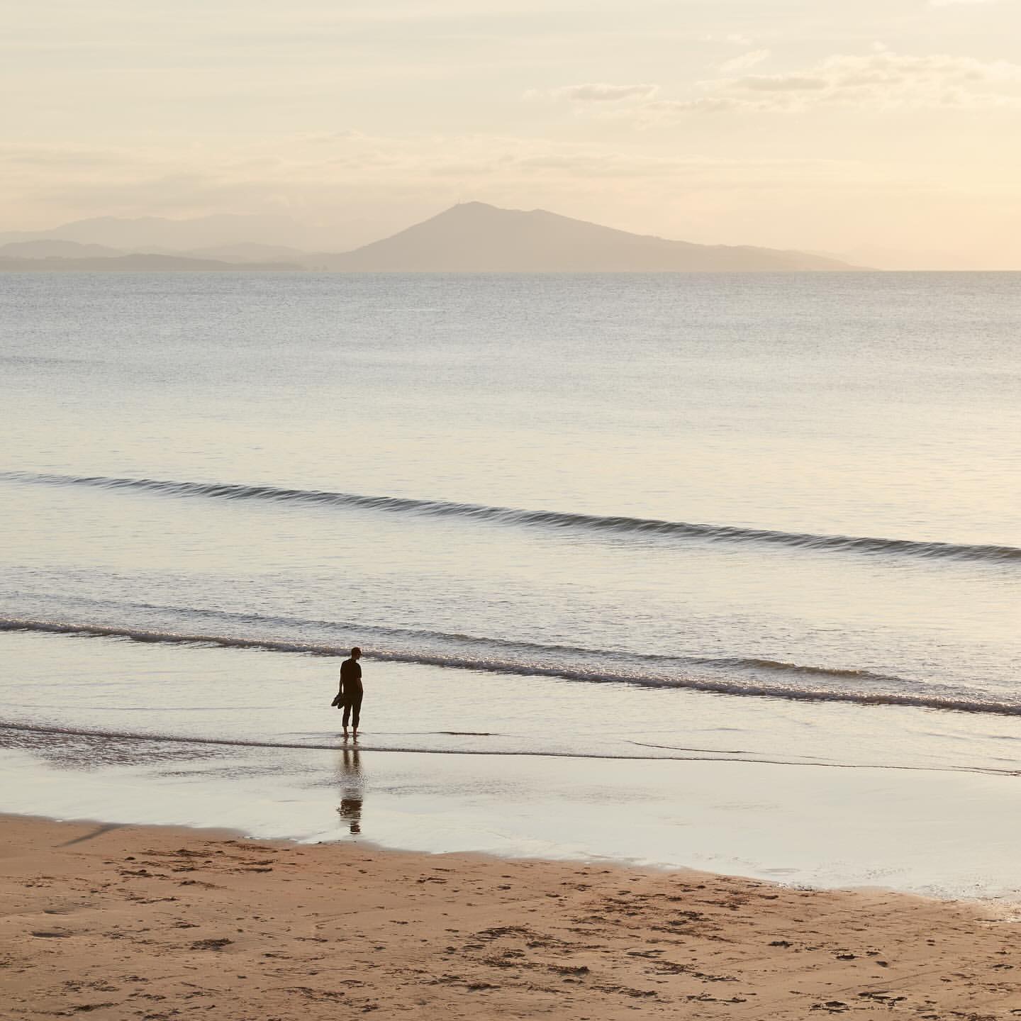 Person standing on a beach with a vast ocean view and a distant mountain under a soft, pastel-colored sky at dusk or dawn.