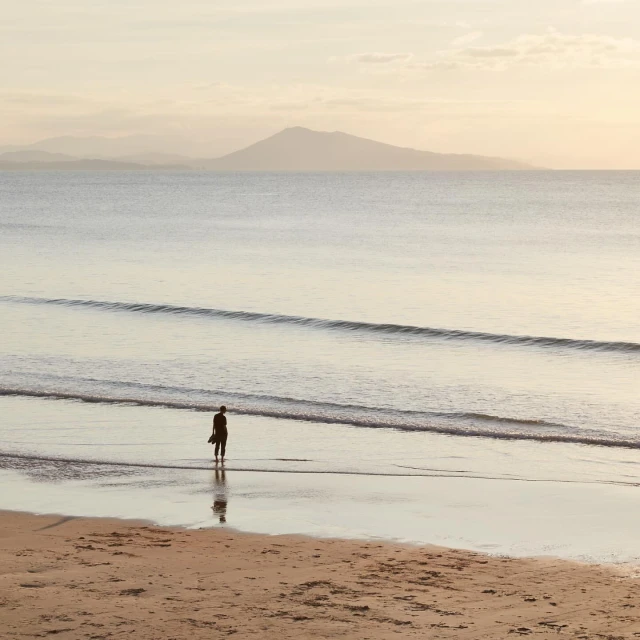 Person standing on a beach with a vast ocean view and a distant mountain under a soft, pastel-colored sky at dusk or dawn.