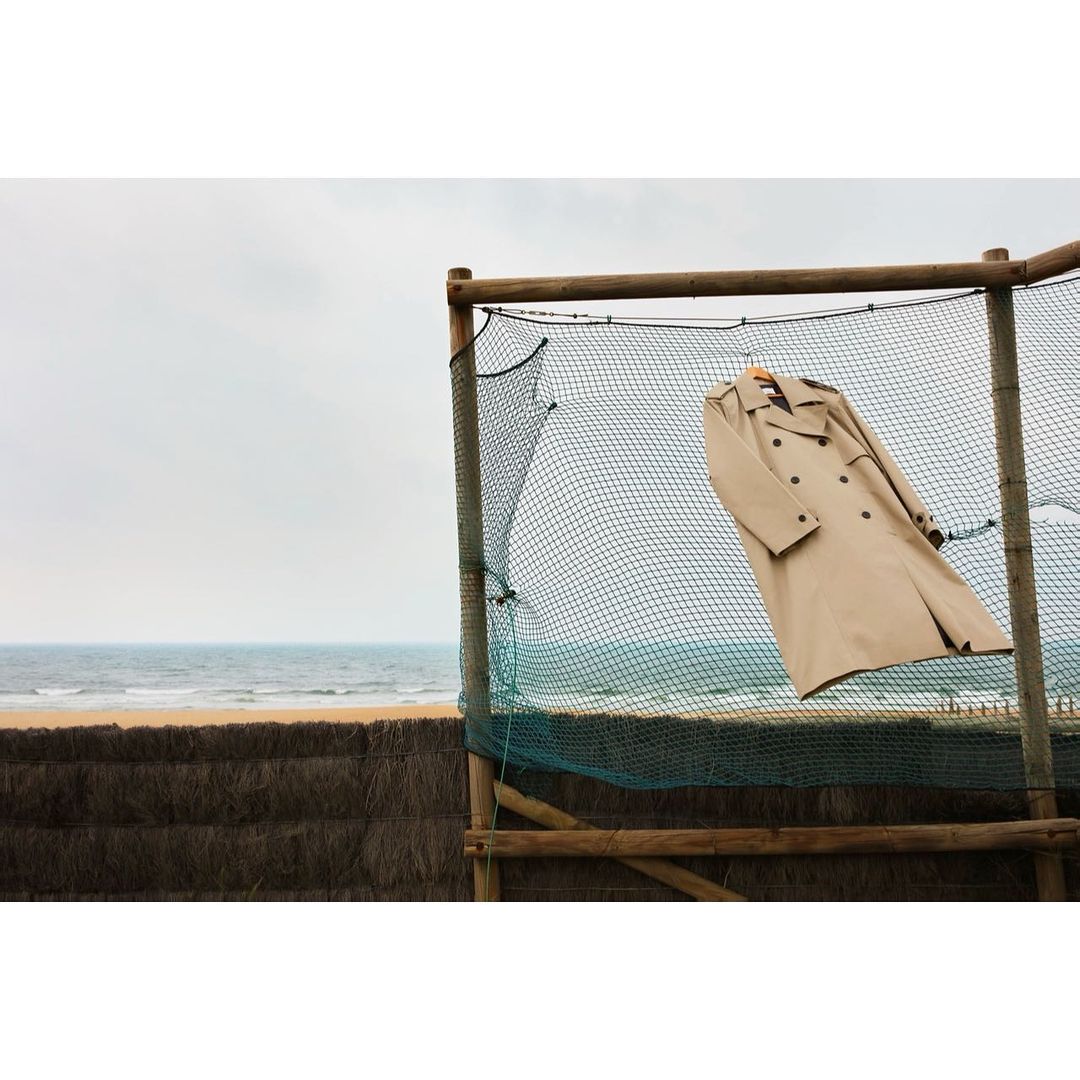 Tan trench coat hanging on a wooden hanger from a fishing net in front of a beach with a cloudy sky.