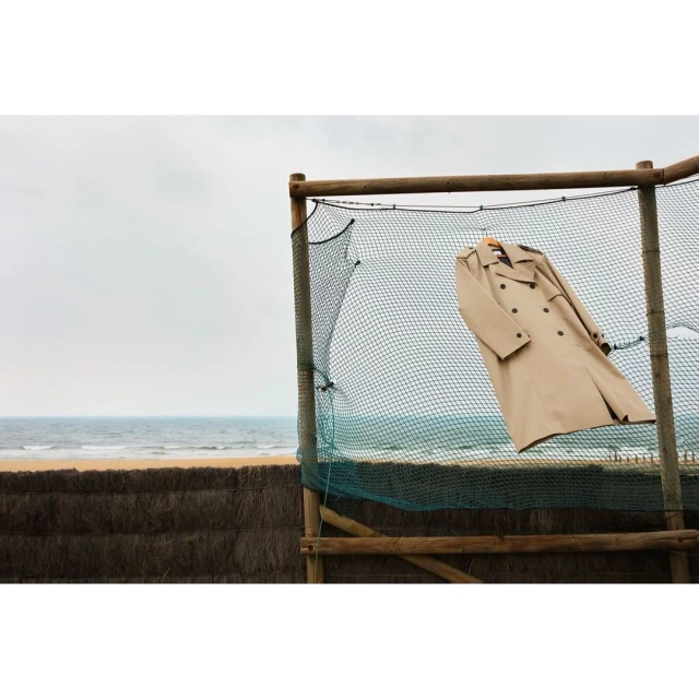 Tan trench coat hanging on a wooden hanger from a fishing net in front of a beach with a cloudy sky.