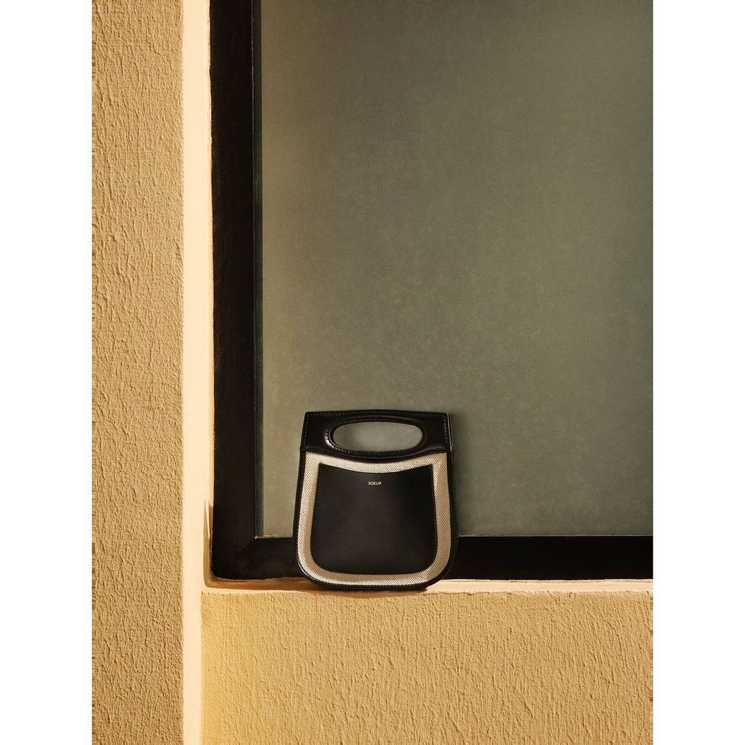 Black tote handbag with a beige trim sitting on a beige ledge in front of a grey window, minimal, modern design, indoor setting.