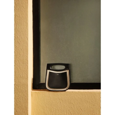 A structured black handbag resting on a textured beige windowsill against a frosted window pane, captured by Roberto Badin.