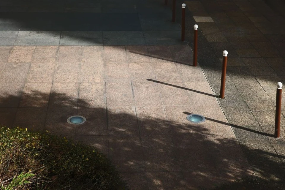 Sidewalk with brown bollards, shadowed plants, and recessed lights. Textured stone paving and geometric shadow patterns create depth.
