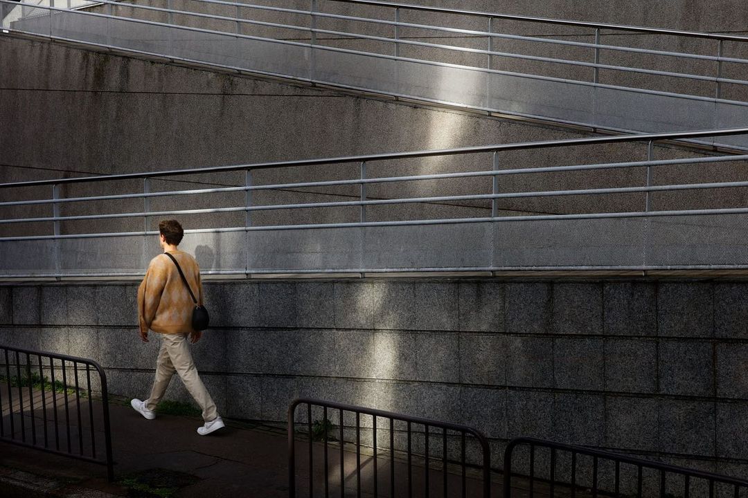 Adult man walking on a pathway beside a concrete wall with metal railings. Wearing casual clothes, light colours and carrying a bag.
