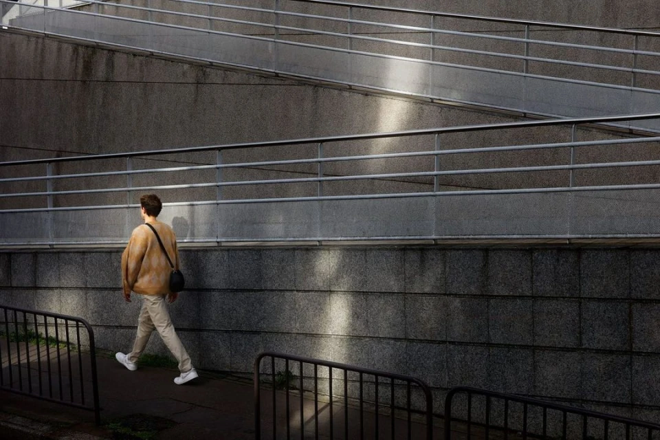 Adult man walking on a pathway beside a concrete wall with metal railings. Wearing casual clothes, light colours and carrying a bag.