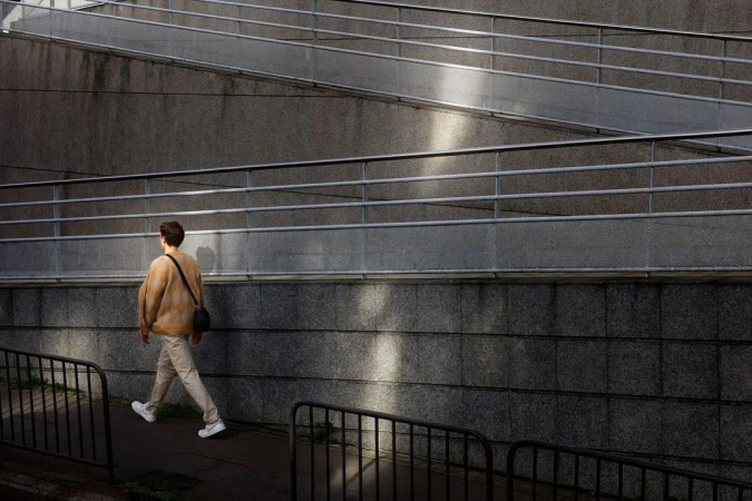 Adult man walking on a pathway beside a concrete wall with metal railings. Wearing casual clothes, light colours and carrying a bag.