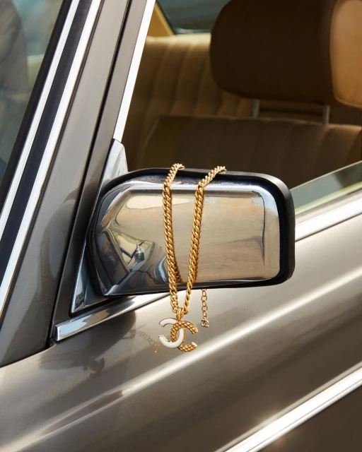 Gold chain necklace with the Chanel logo draped over the side mirror of a gray vehicle with tan interior visible through the window.
