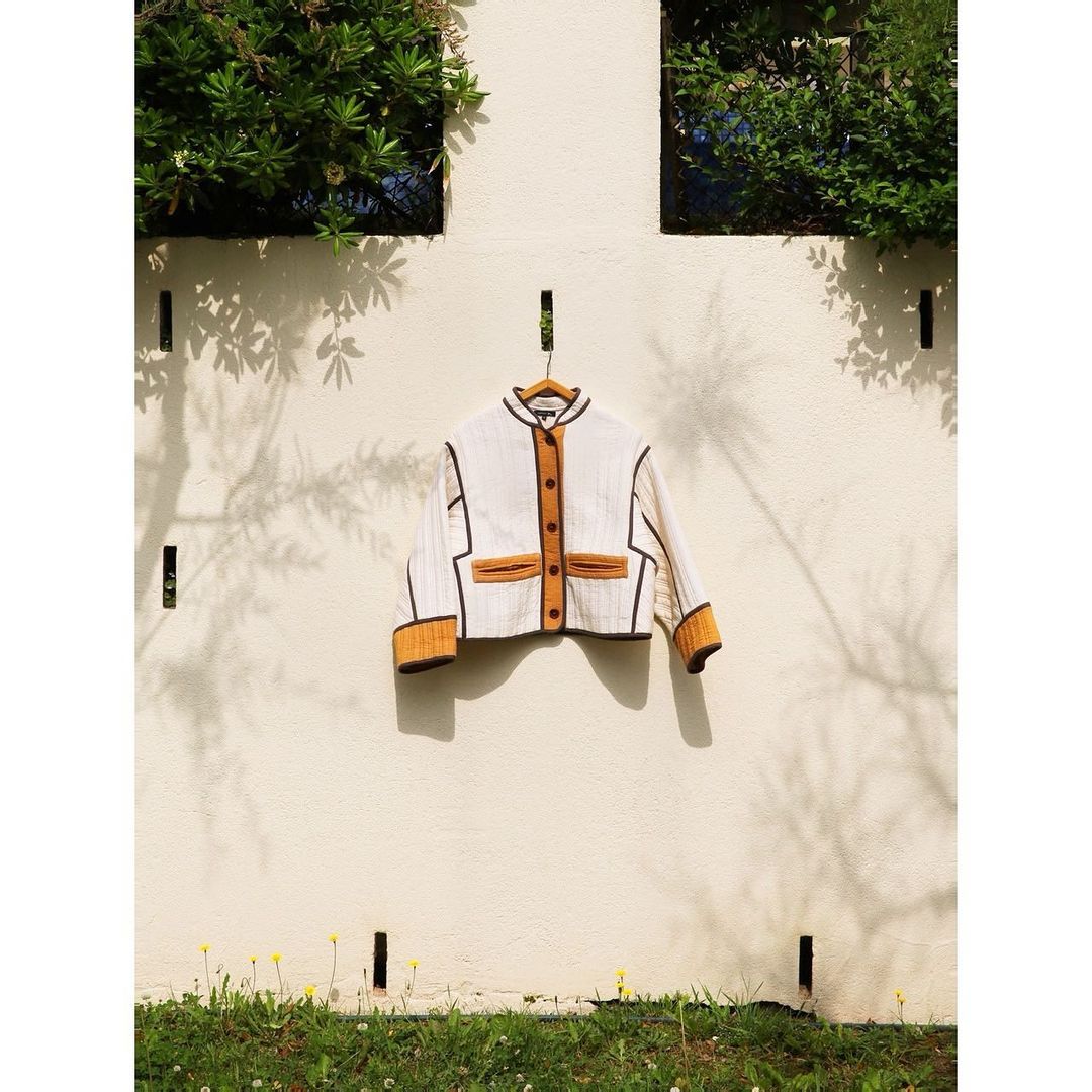 Jacket with mustard and brown accents hung outdoors on a wooden hanger against a white wall, framed by green foliage.