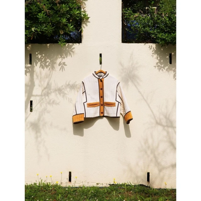 Jacket with mustard and brown accents hung outdoors on a wooden hanger against a white wall, framed by green foliage.