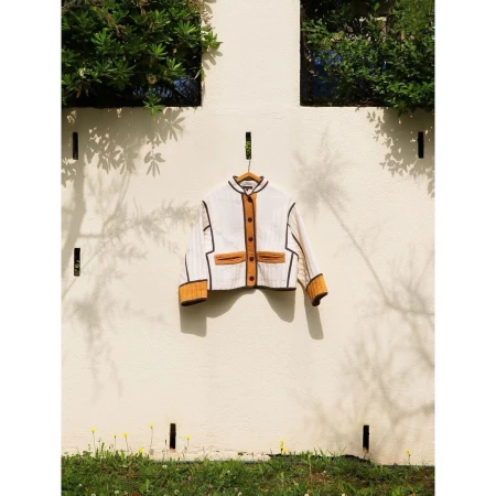 Jacket with mustard and brown accents hung outdoors on a wooden hanger against a white wall, framed by green foliage.
