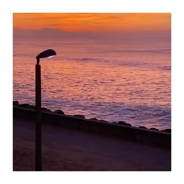 Tranquil coastal scene at sunset features a solitary lamppost overlooking the sea under a gradient orange sky.
