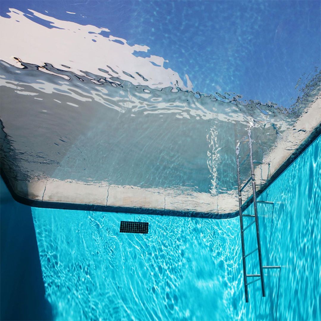 Underwater view of a swimming pool with clear turquoise water, a metal ladder, and a section of the pool's white side.