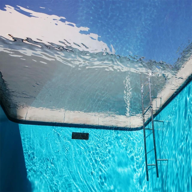 Underwater view of a swimming pool with clear turquoise water, a metal ladder, and a section of the pool's white side.