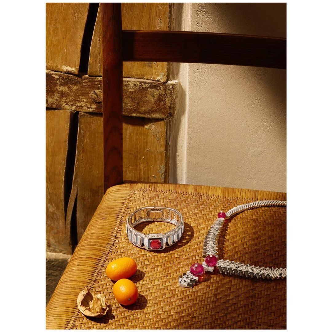 Still life photo of a diamond bracelet and necklace with red gemstones, arranged on a woven chair with tangerines and a walnut.