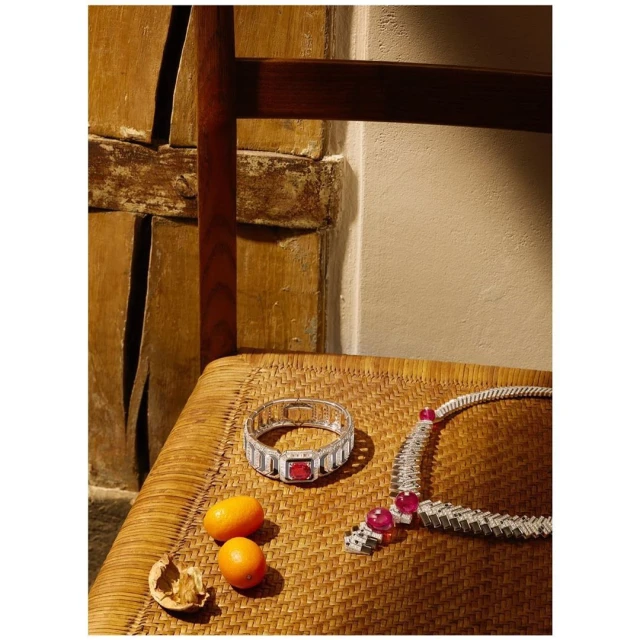 Still life photo of a diamond bracelet and necklace with red gemstones, arranged on a woven chair with tangerines and a walnut.