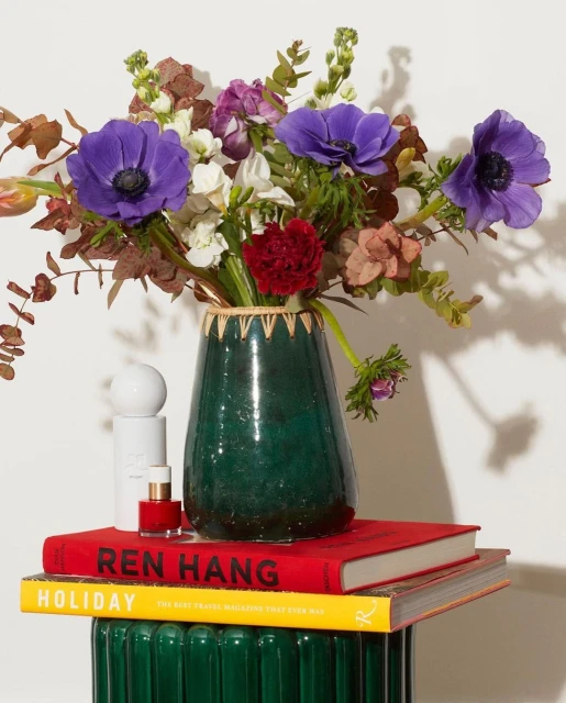 Floral arrangement with purple anemones in a green vase atop stacked books and a ridged green pedestal against a white background.
