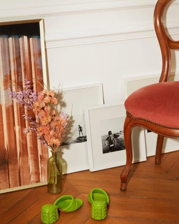 Green heeled mules on a wood floor beside a vase of flowers, framed artworks, and a wooden chair with a red seat.