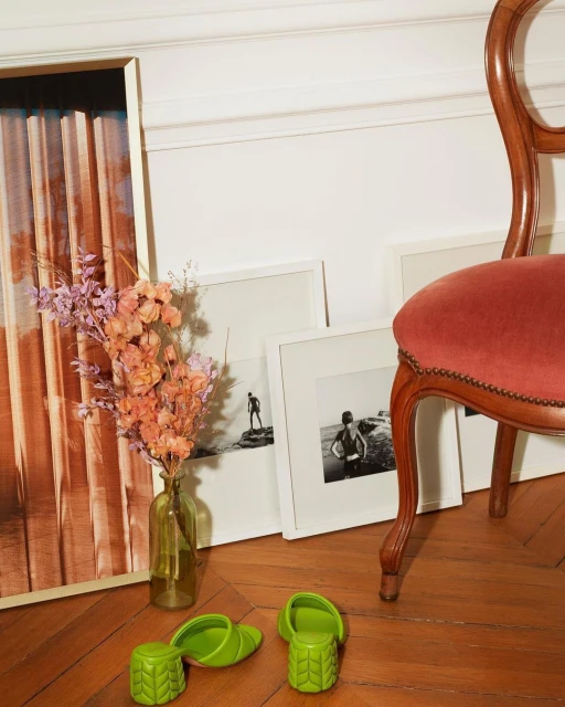 A pair of green platform shoes sits on a wood floor next to framed artwork and a vase of flowers, beside a wood and velvet chair.