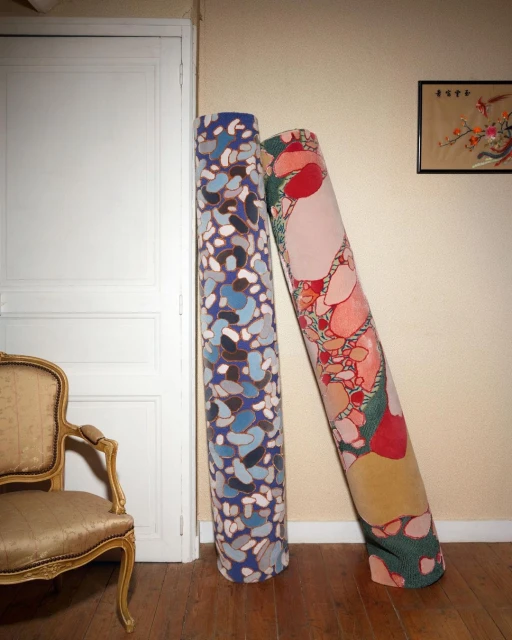 Two rolls of patterned textile lean against a wall in a room with vintage furniture and a door, showcasing interior design and textile art.