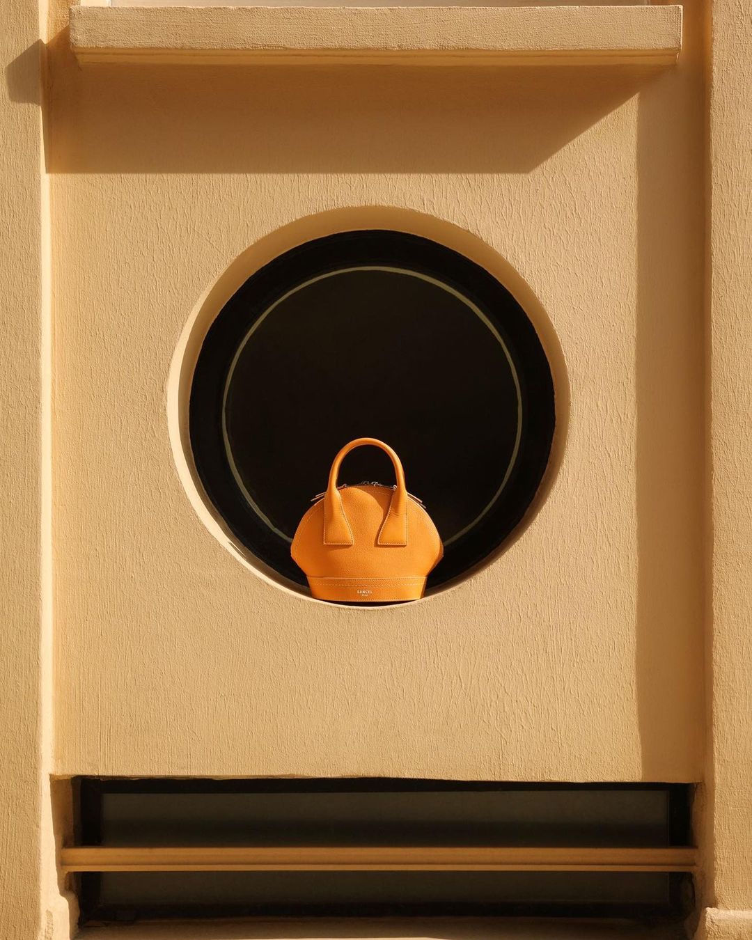 A handbag is positioned in a circular opening within a light beige wall, creating a minimalist, architectural fashion product shot.
