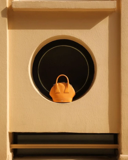 A handbag is positioned in a circular opening within a light beige wall, creating a minimalist, architectural fashion product shot.