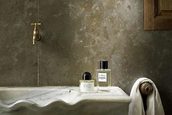 Two Chanel perfume bottles are displayed on a white marble vanity next to a decorative brass faucet and a white towel.
