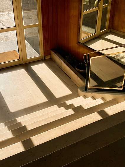 Indoor architectural shot featuring stairs, sunlight patterns, a glass door, wooden paneling, and minimalist decor creating geometric shadows.