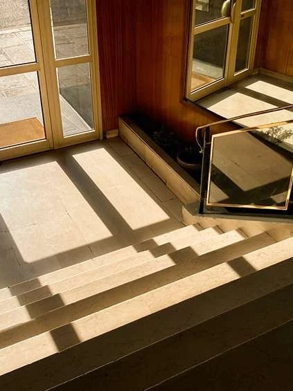 Indoor architectural shot featuring stairs, sunlight patterns, a glass door, wooden paneling, and minimalist decor creating geometric shadows.