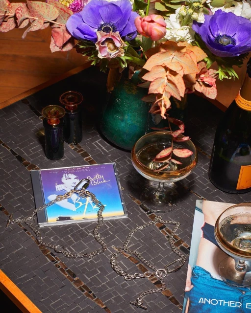 Still life composition featuring a floral arrangement, champagne glasses, champagne bottle, jewelry, and a CD on a textured surface.