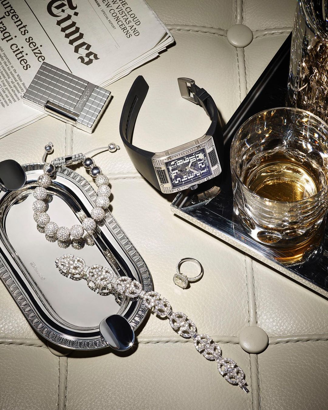 Wristwatch, jewelry, and barware are arranged on a tufted surface with a newspaper, lighter, and silver trays under studio lighting.